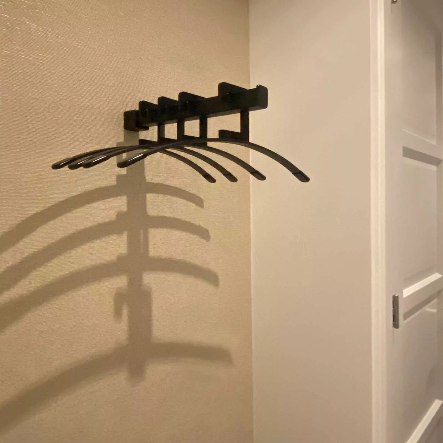 Wandgarderobe Imora quadratisch schwarz an der Wand neben einer Tür mit leeren Kleiderbügeln und Schatten an der Wand montiert.